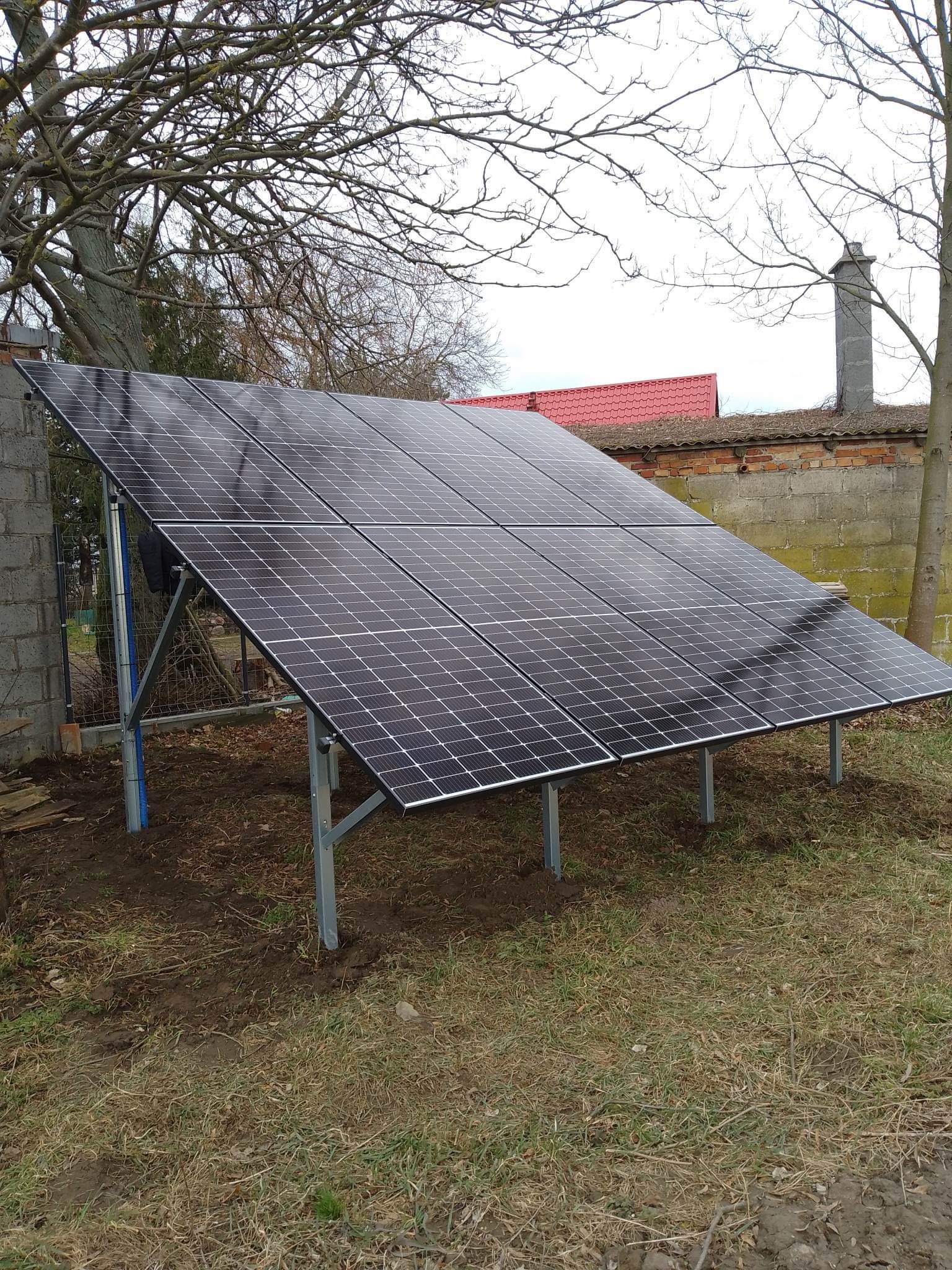 Panel słoneczny zamontowany na metalowej konstrukcji na gruncie, widoczne przewody w niebieskiej rurce ochronnej, w tle mur z czerwonej cegły i komin.