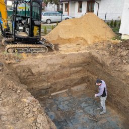 Solid-Tech Lucjan Golonka - Głęboki wykop z równymi ścianami, na dnie którego mężczyzna wyrównuje grabiami powierzchnię; obok stoi mini koparka, a w tle widać dom i stertę ziemi.