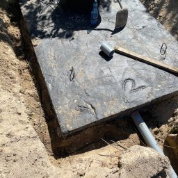 Solid-Tech Lucjan Golonka - Betonowy zbiornik z pokrywą, częściowo zakopany w ziemi, z widocznymi rurami odpływowymi i narzędziami na pokrywie, takimi jak łopata i poziomica, oraz butelka wody mineralnej