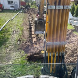 Solid-Tech Lucjan Golonka - Żółta koparka z łyżką w wykopie, widok z kabiny operatora, rury drenażowe leżące obok wykopu, budynki w tle.