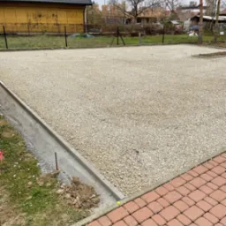 Utwardzony teren drobnym kruszywem z betonową obwódką, widoczny fragment chodnika z kostki brukowej, w tle budynek i ogrodzenie z siatki.