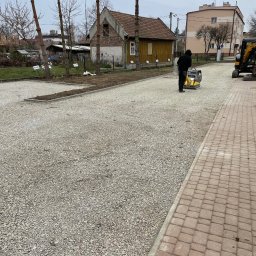 Solid-Tech Lucjan Golonka - Pracownik w ciemnej odzieży zagęszcza podłoże z kruszywa na nowo powstałym parkingu przy użyciu zagęszczarki wibracyjnej, widoczny fragment chodnika z kostki brukowej, w tle budynek mieszkalny...