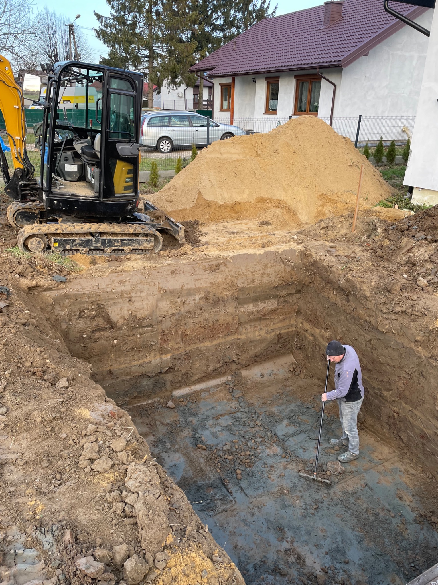 Głęboki wykop z równymi ścianami, na dnie którego mężczyzna wyrównuje grabiami powierzchnię; obok stoi mini koparka, a w tle widać dom i stertę ziemi.