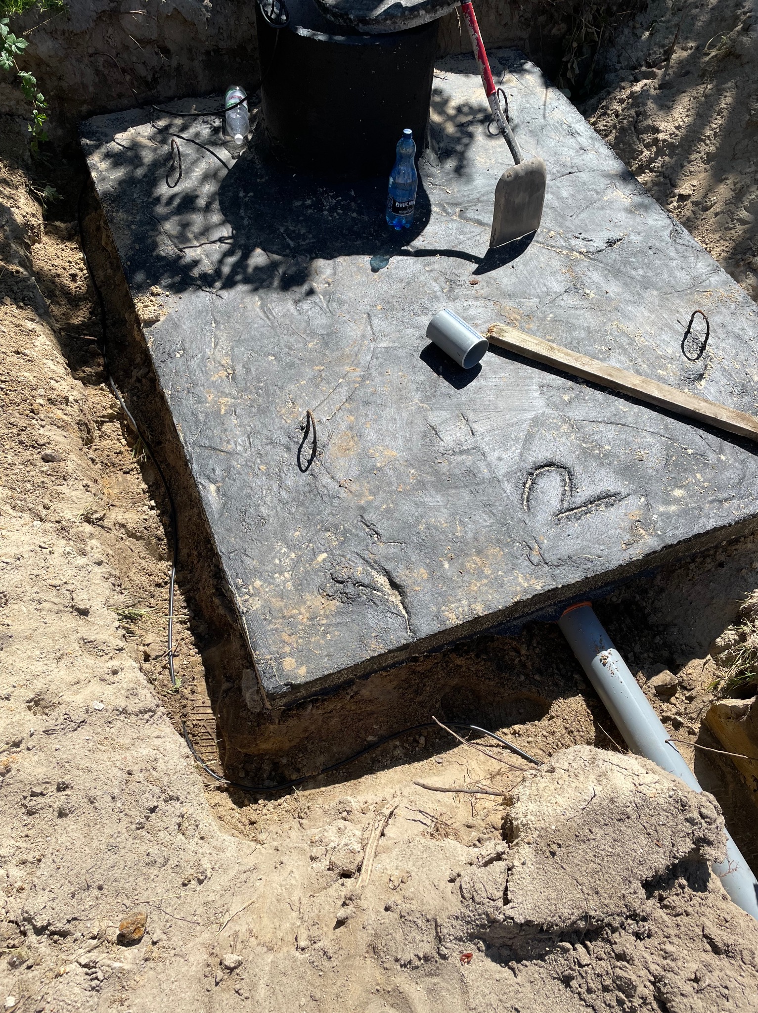 Betonowy zbiornik z pokrywą, częściowo zakopany w ziemi, z widocznymi rurami odpływowymi i narzędziami na pokrywie, takimi jak łopata i poziomica, oraz butelka wody mineralnej