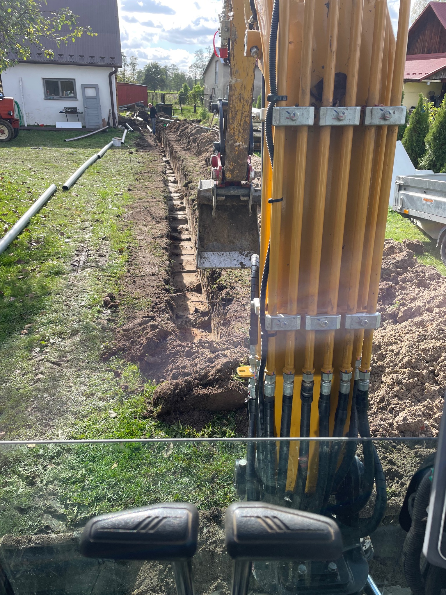 Żółta koparka z łyżką w wykopie, widok z kabiny operatora, rury drenażowe leżące obok wykopu, budynki w tle.