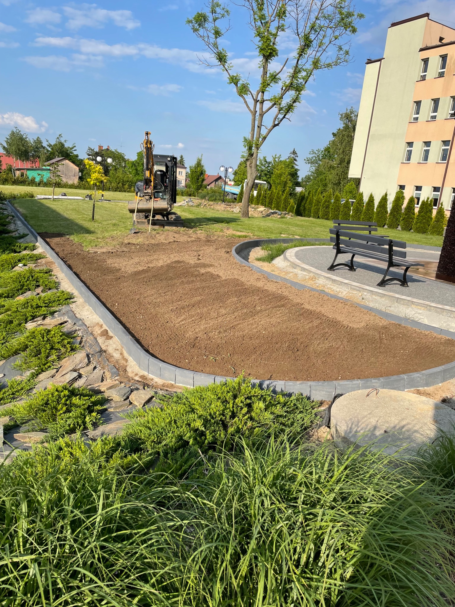 Przygotowany teren pod trawnik z obrzeżami, widoczny mini-koparka na trawie w tle, ławka parkowa i budynek.