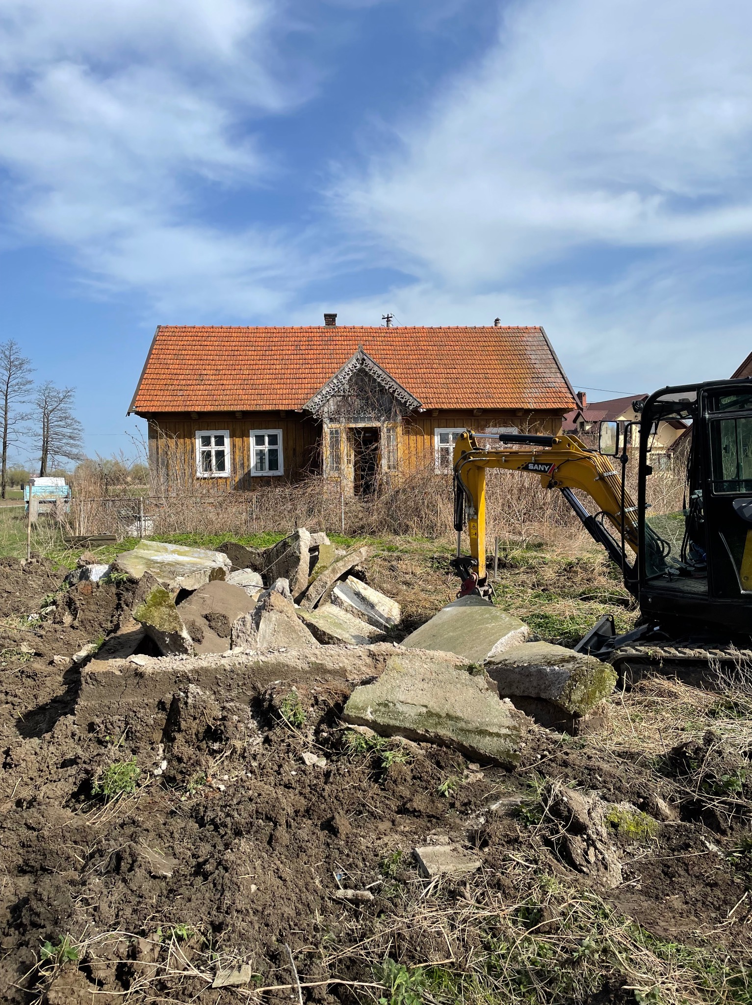 Żółty minikoparka SANY na tle starego, drewnianego domu z pomarańczową dachówką, usuwająca gruz betonowy z terenu.