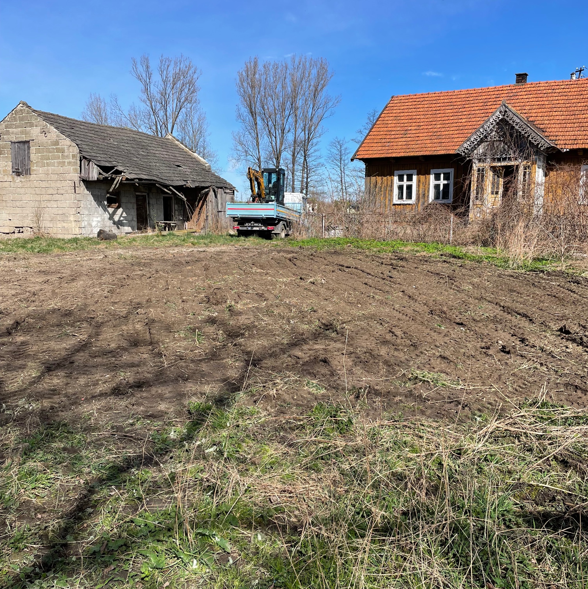 Przygotowany teren pod budowę z widocznymi śladami po maszynach, w tle stary budynek gospodarczy z cegły i drewniany dom z pomarańczową dachówką, na podwórku stoi mała koparka na lawecie samochodu...