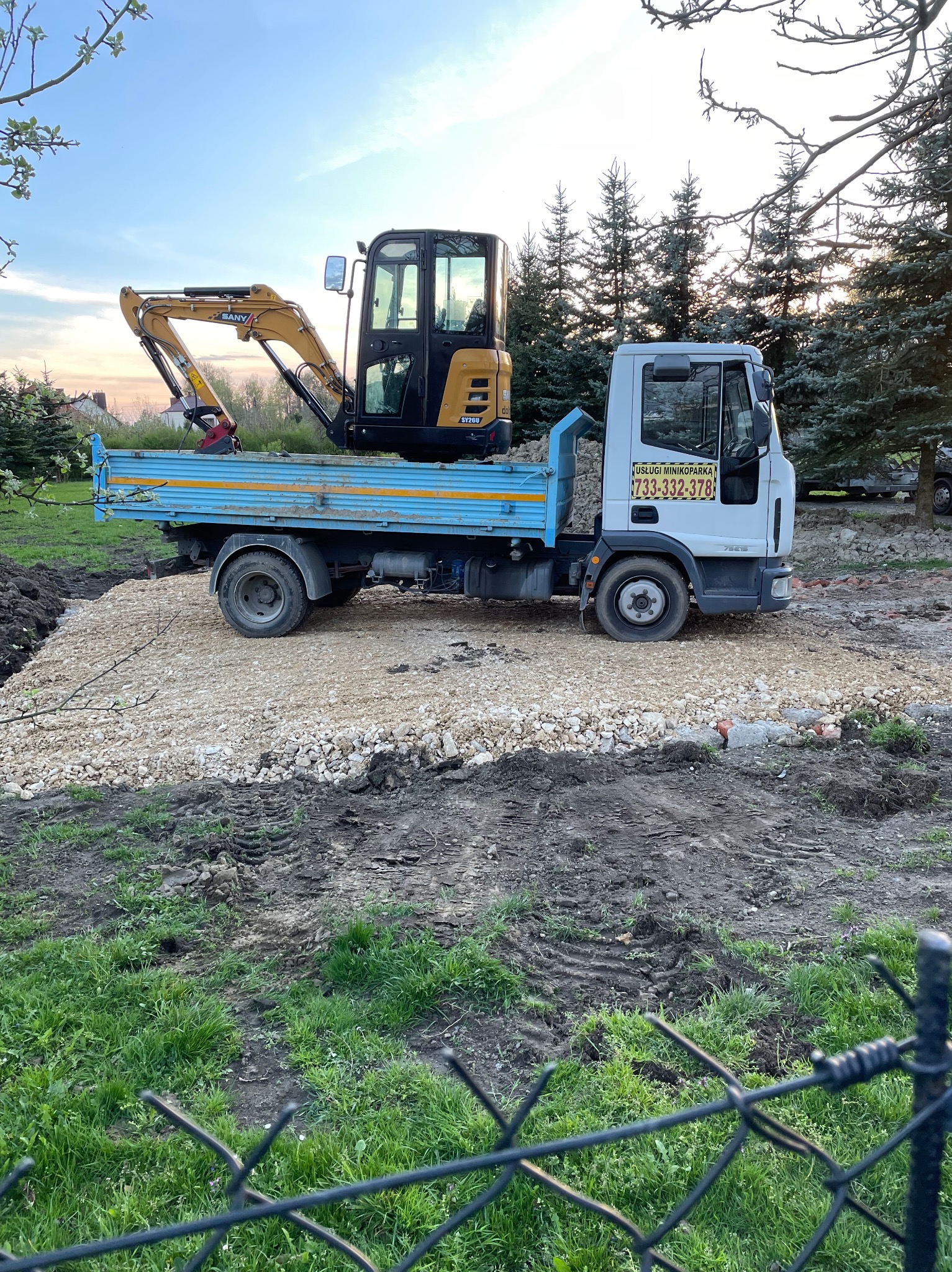 Niebieska ciężarówka z żółtą mini koparką SANY na pace, stojąca na świeżo utwardzonej powierzchni z białego kruszywa, widok zza ogrodzenia.