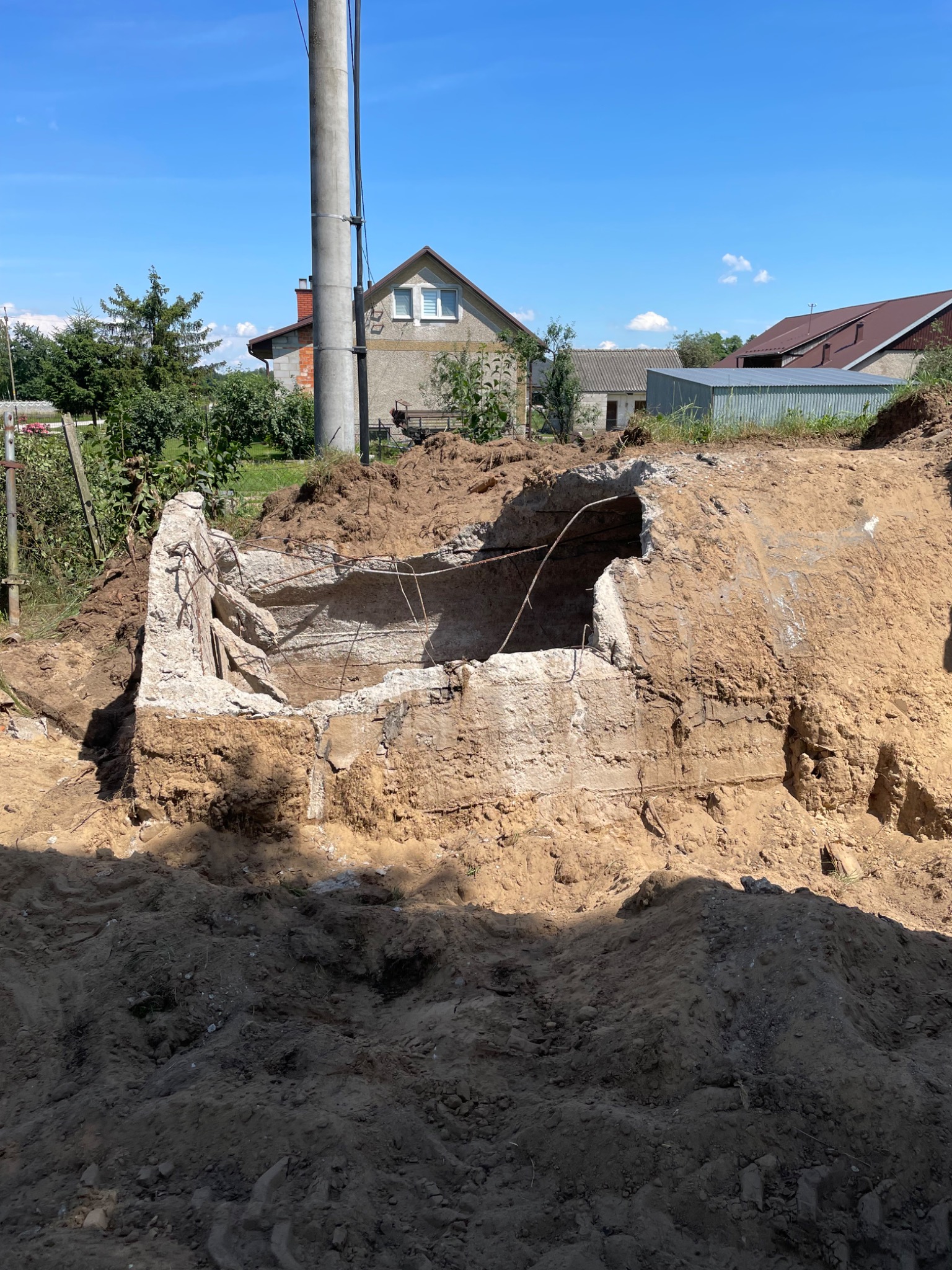 Zniszczona betonowa konstrukcja z wystającymi prętami zbrojeniowymi, częściowo zasypana ziemią, na tle krajobrazu wiejskiego z domami i słupem elektrycznym.