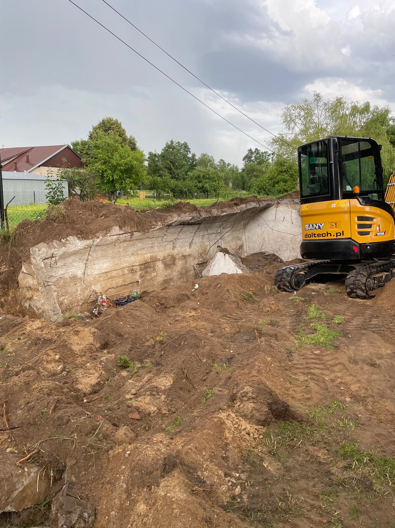 Żółta minikoparka SANY stoi obok wykopu z odsłoniętymi betonowymi ścianami z widocznym zbrojeniem, częściowo zasypanymi ziemią, w tle zieleń i pochmurne niebo.
