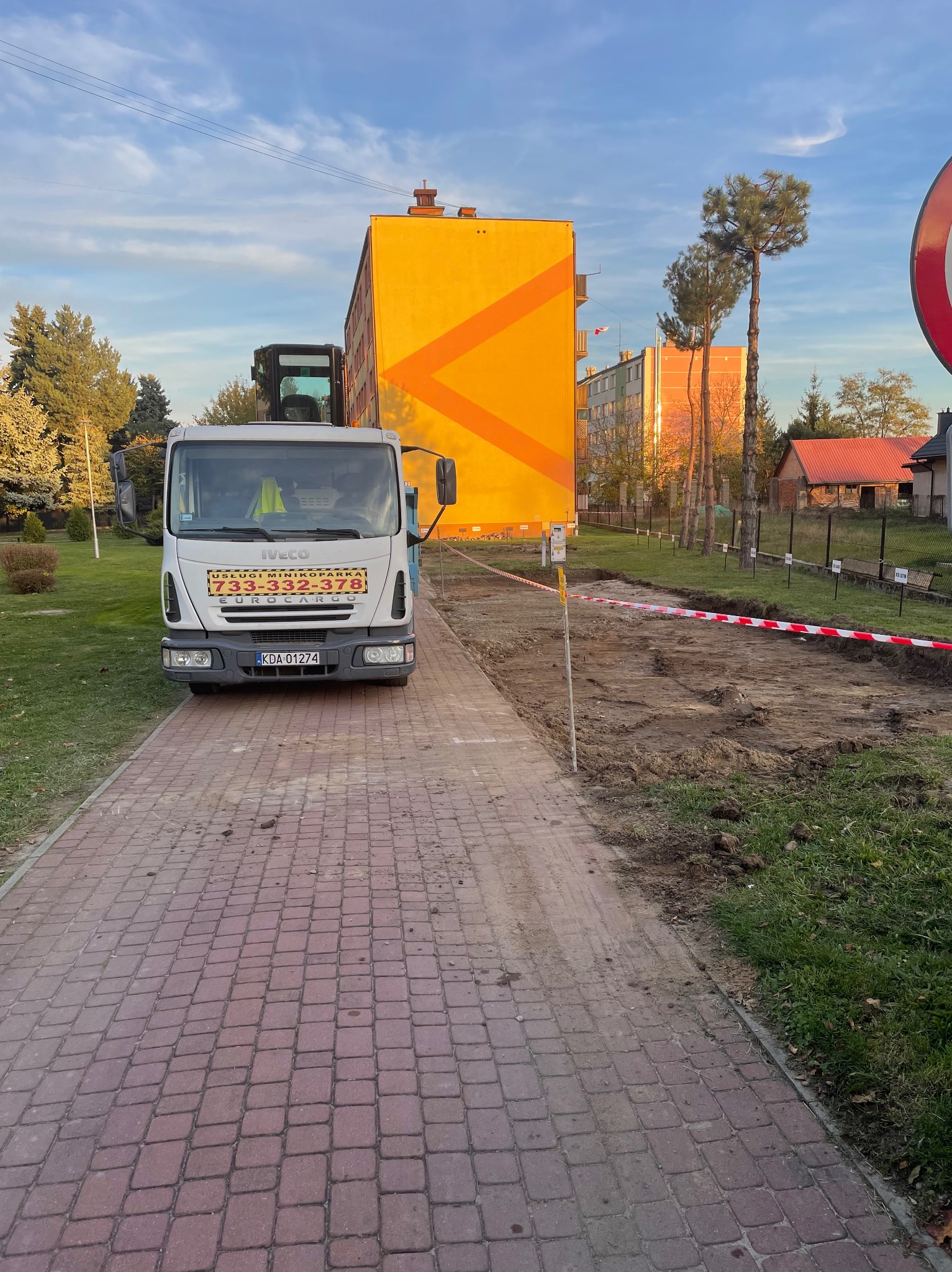 Ciężarówka firmy budowlanej parkuje obok wykopu zabezpieczonego taśmą ostrzegawczą, w tle budynek mieszkalny z pomarańczowym akcentem.