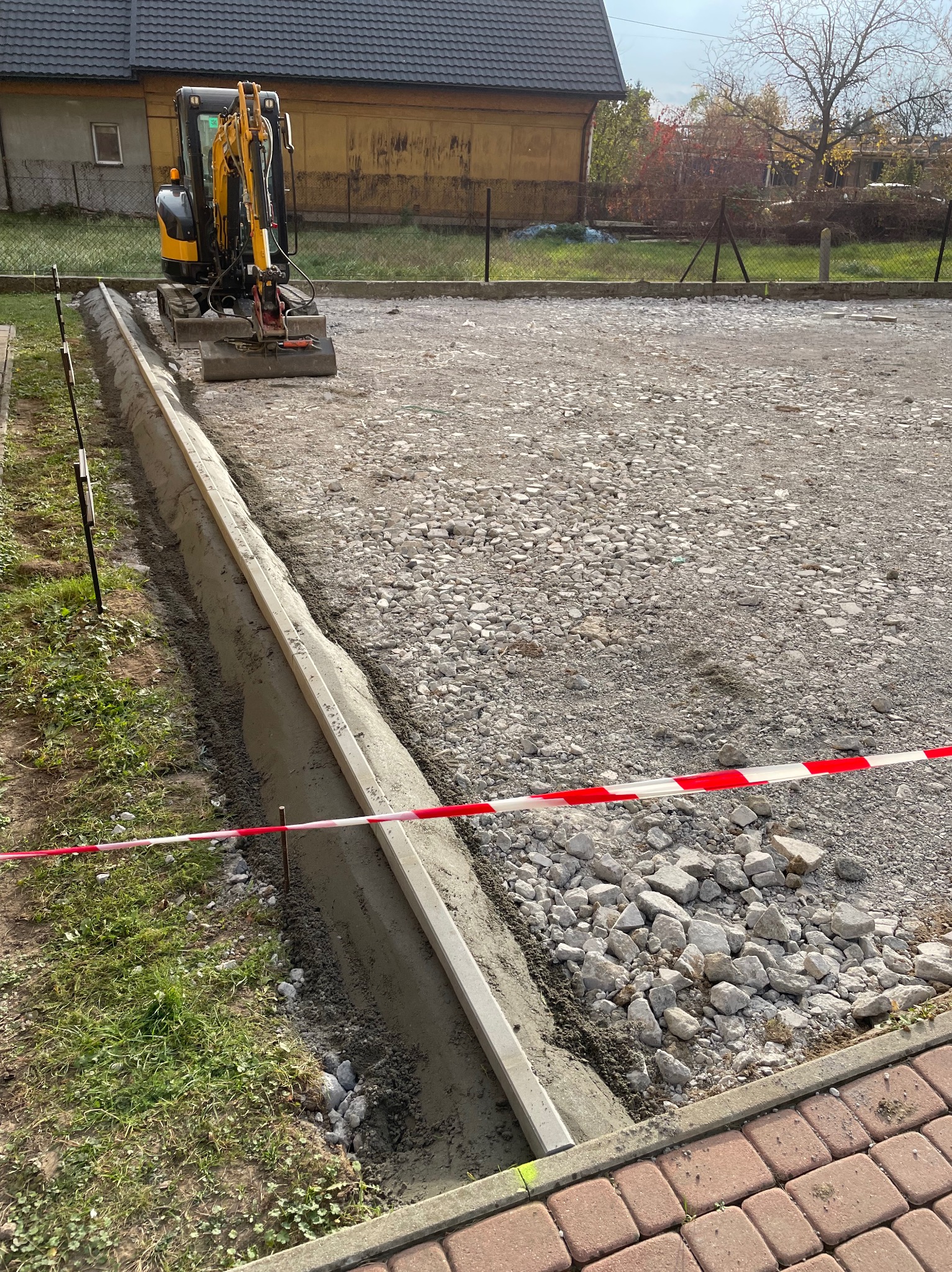 Przygotowanie podłoża pod drogę: mini koparka, warstwa gruzu, świeżo wylany betonowy obrzeże z listwą poziomującą, taśma ostrzegawcza, widok z góry w słoneczny dzień.