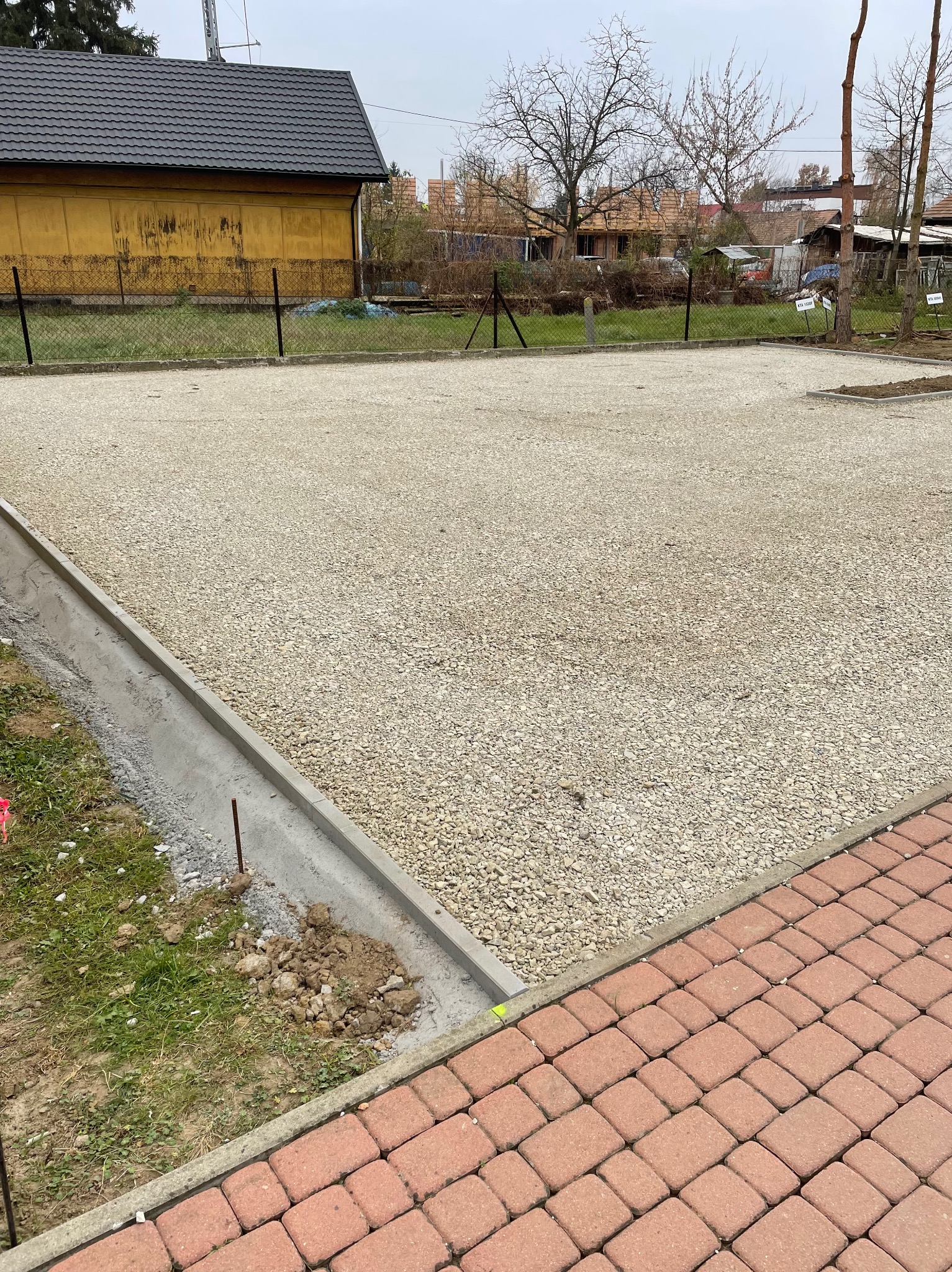 Utwardzony teren drobnym kruszywem z betonową obwódką, widoczny fragment chodnika z kostki brukowej, w tle budynek i ogrodzenie z siatki.