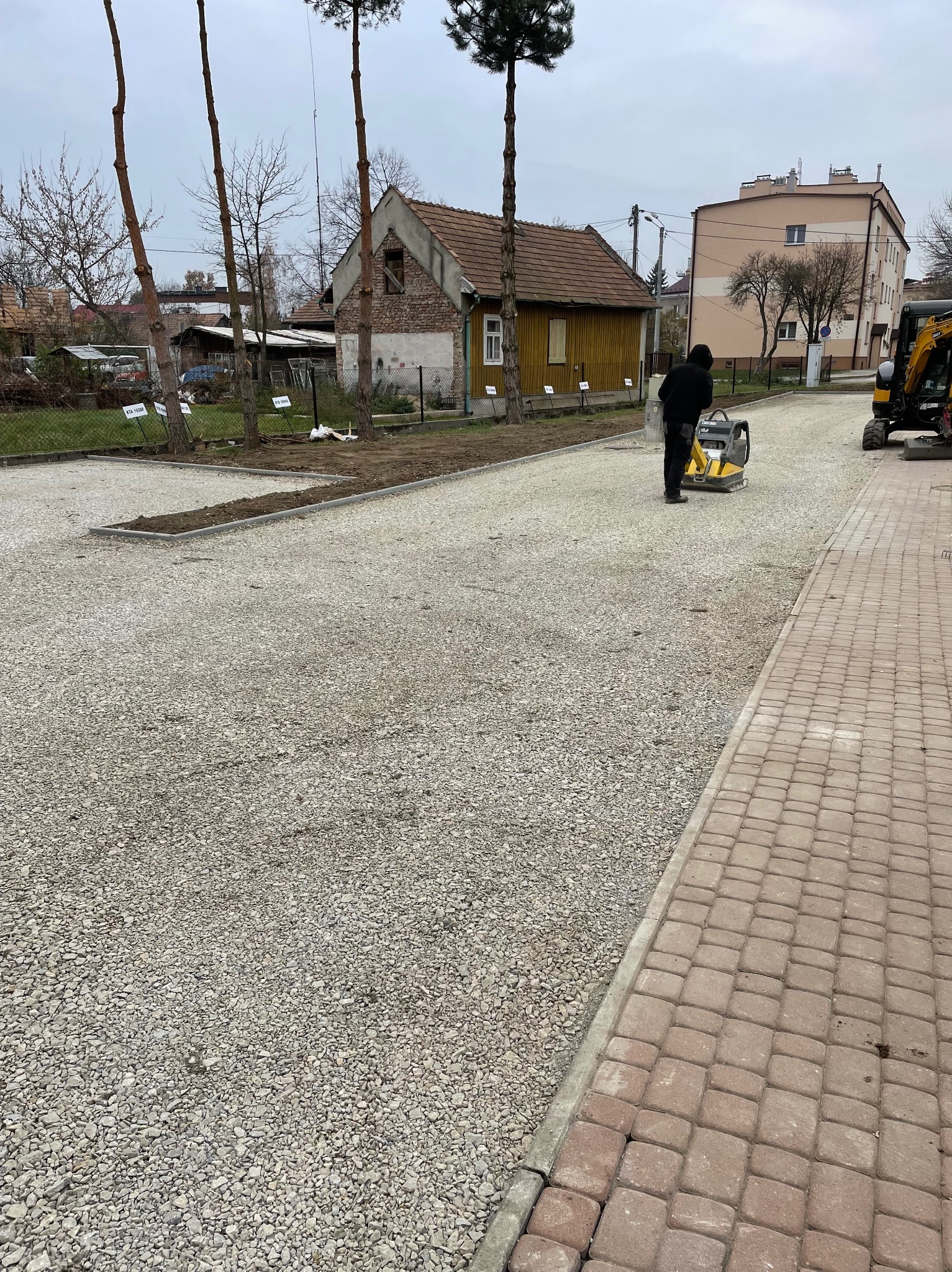 Pracownik w ciemnej odzieży zagęszcza podłoże z kruszywa na nowo powstałym parkingu przy użyciu zagęszczarki wibracyjnej, widoczny fragment chodnika z kostki brukowej, w tle budynek mieszkalny...
