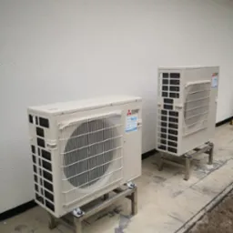 Dwa beżowe agregaty klimatyzacji Mitsubishi Electric zamontowane na metalowych podstawach, ustawione na szarym betonowym podłożu przy jasnej ścianie budynku.