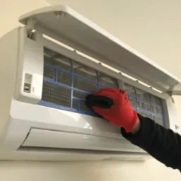 Otwarty panel klimatyzatora ściennego, widoczny filtr siatkowy, dłoń w czerwonej rękawicy roboczej z czarnymi wzmocnieniami, podczas czyszczenia lub wymiany filtra.