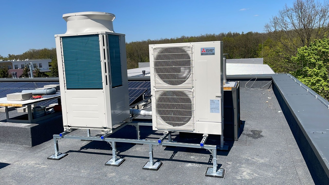 Dwie jednostki zewnętrzne klimatyzacji Mitsubishi Electric na dachu budynku, zamontowane na metalowej konstrukcji wsporczej z regulowanymi nóżkami, widoczne panele słoneczne w tle.
