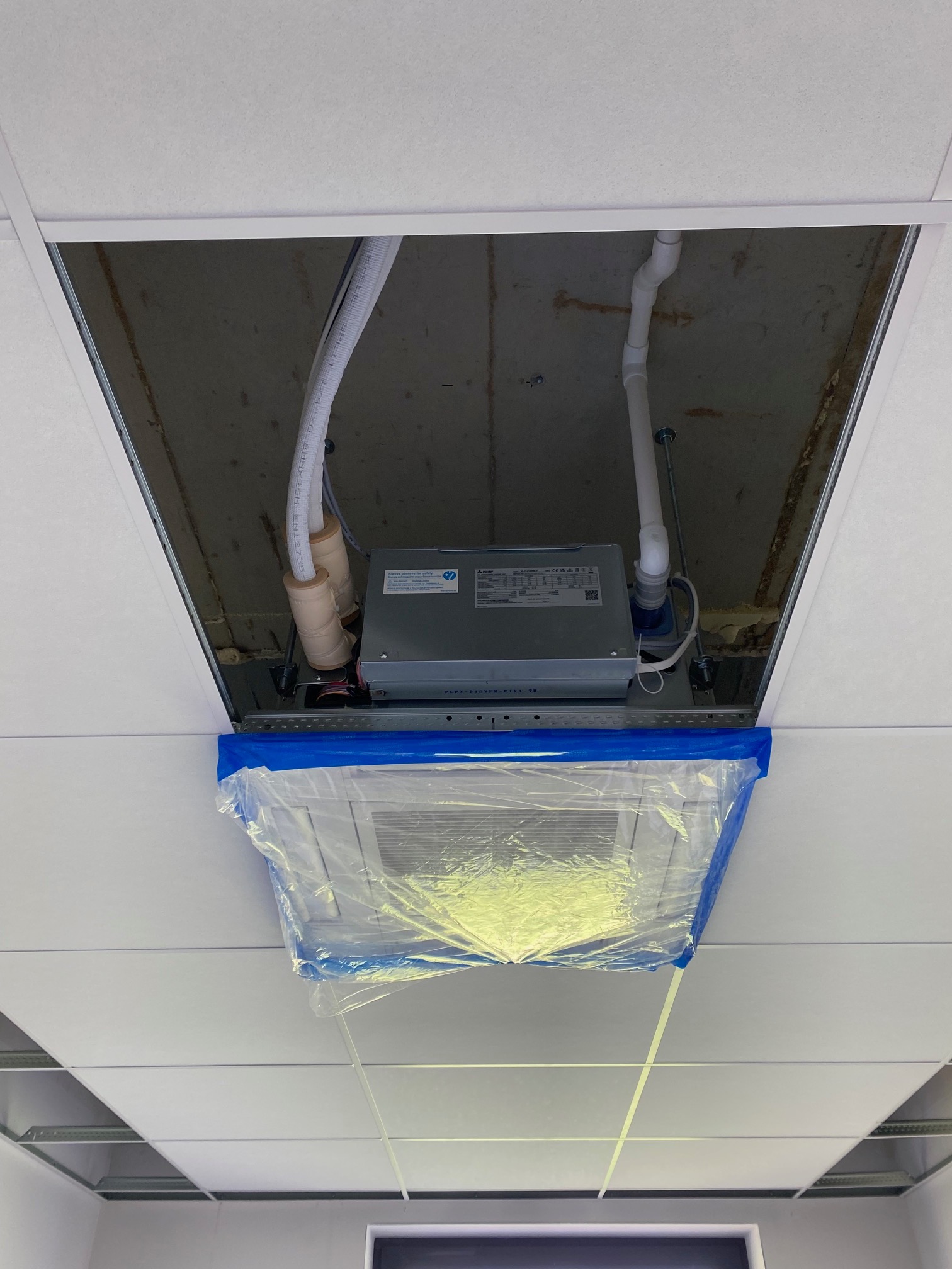 Kaseton sufitowy z zamontowaną jednostką wewnętrzną klimatyzacji zabezpieczoną folią i taśmą malarską, widoczne rury i instalacja w przestrzeni nad sufitem podwieszanym.