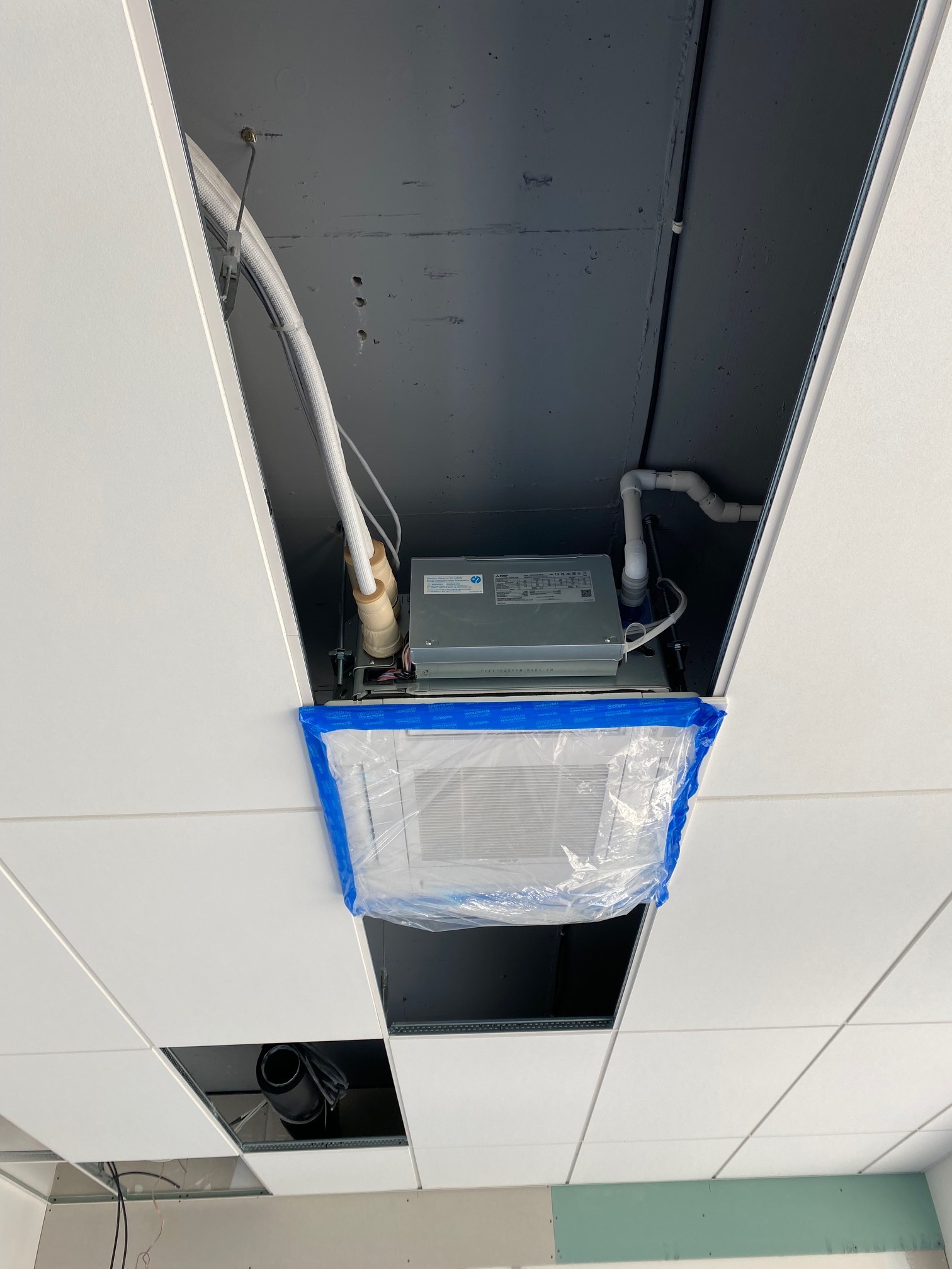 Widok od dołu na montowany klimakonwektor kasetonowy w suficie podwieszanym, zabezpieczony folią ochronną. Widoczne białe panele sufitowe, czarne elementy konstrukcyjne nad sufitem...