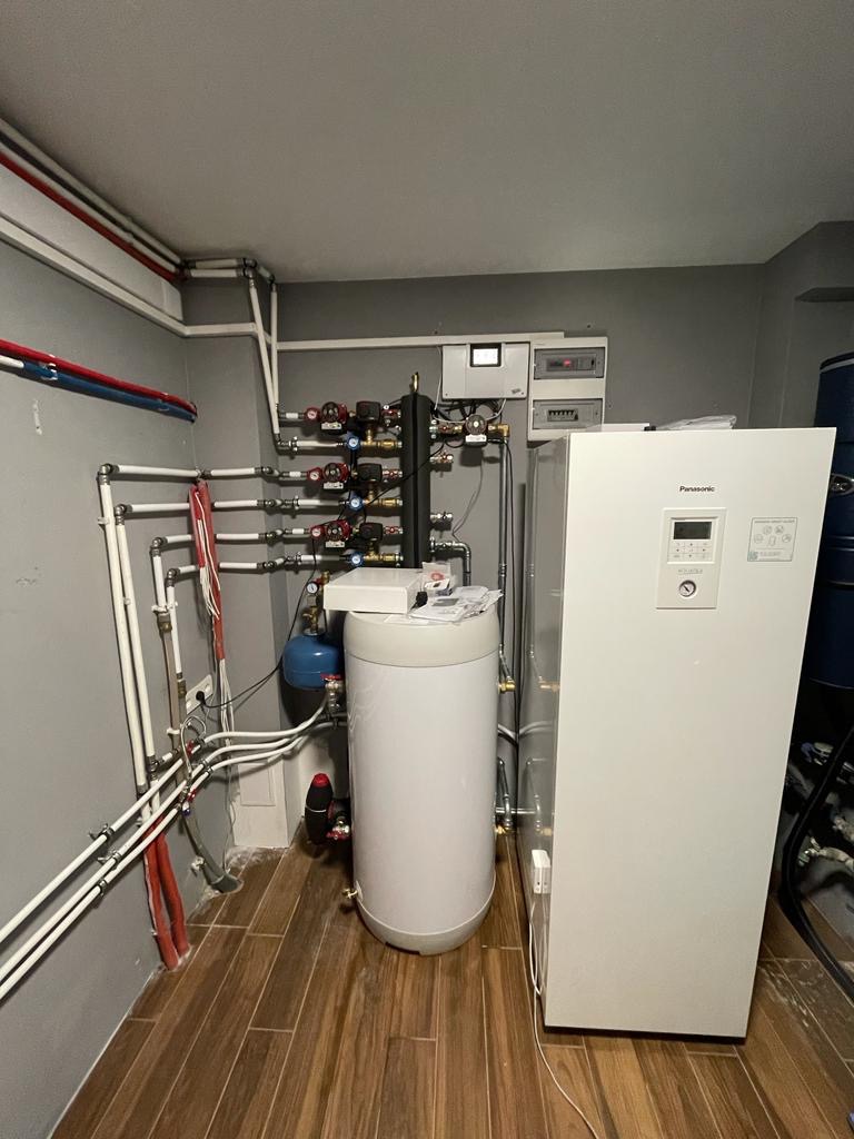 Zestaw urządzeń w kotłowni: biały zasobnik ciepłej wody, pompa ciepła Panasonic, instalacja rurowa z zaworami, licznik energii elektrycznej na szarej ścianie i drewnopodobna podłoga.