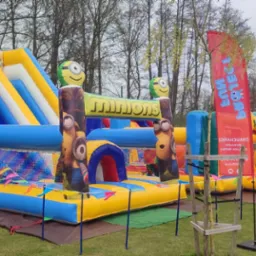 Duży, kolorowy dmuchany plac zabaw z motywem Minionków, zjeżdżalnie i przeszkody, ustawiony na trawiastym terenie zewnętrznym w pobliżu drzew i drewnianej altany.