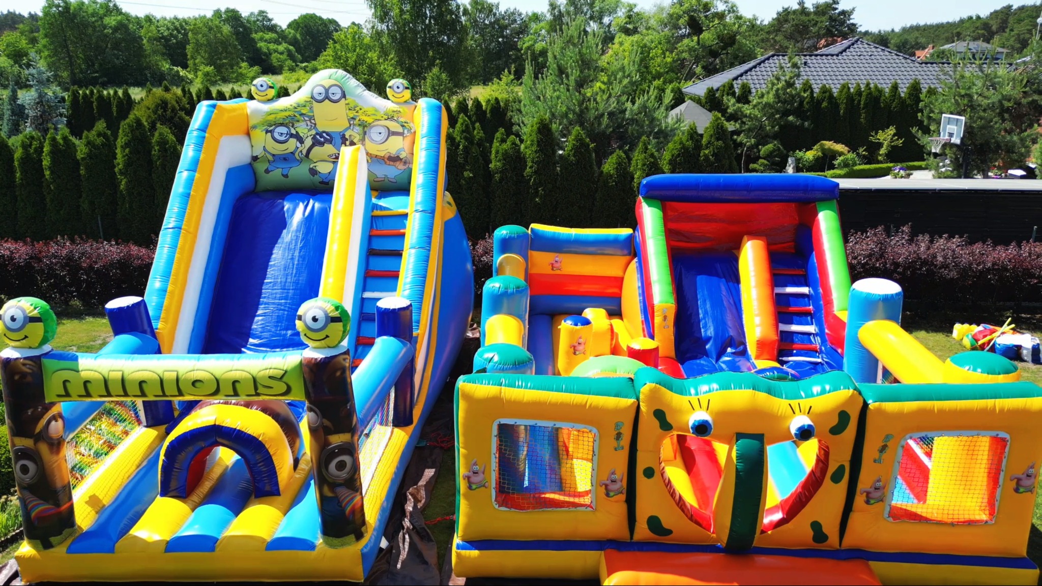 Dwa kolorowe, dmuchane zamki z motywami Minionków i SpongeBoba, ustawione na trawniku przed domem z zielonymi drzewami w tle.