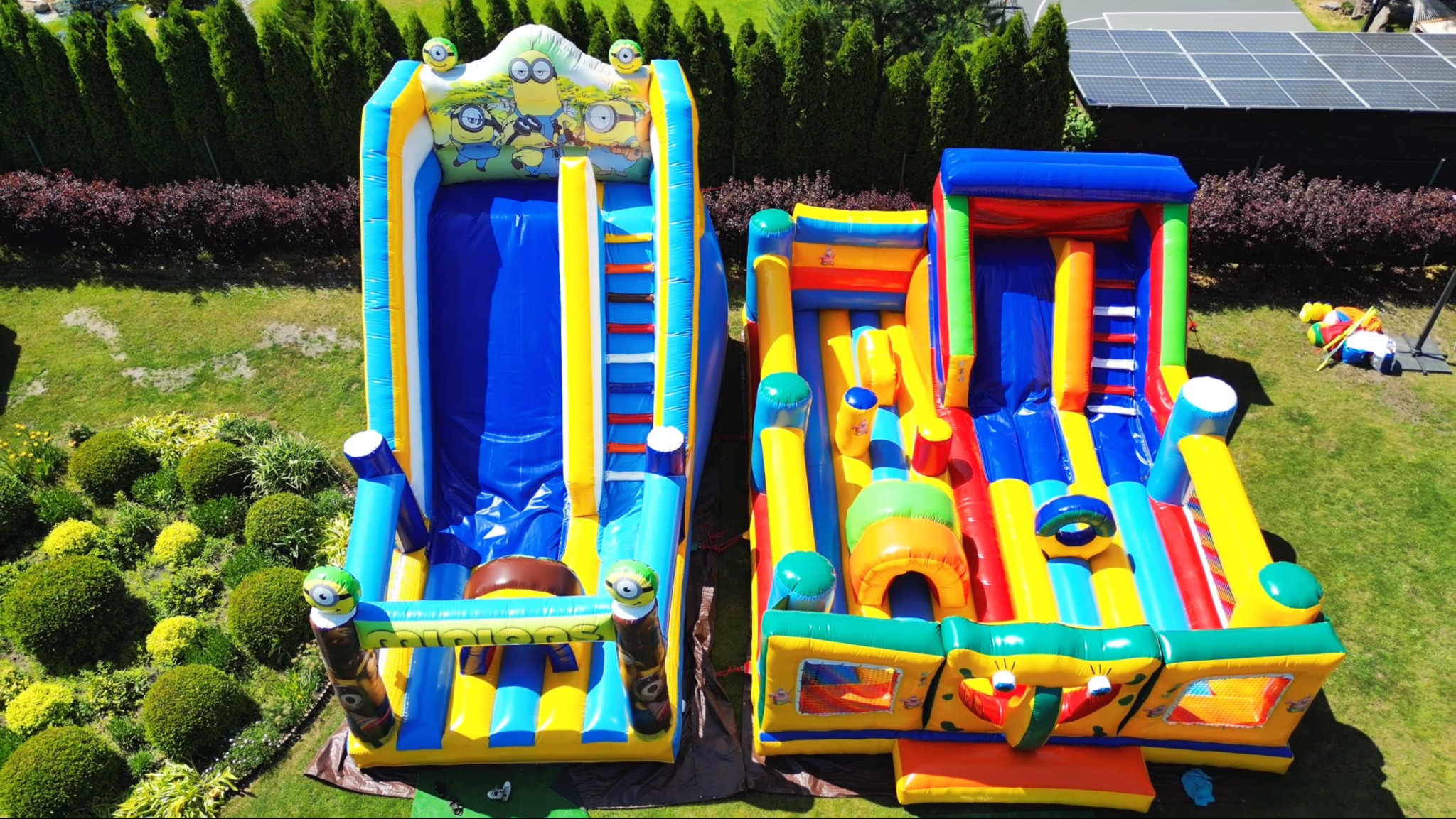 Dwa kolorowe dmuchańce na trawniku, jeden ze zjeżdżalnią z motywem Minionków, drugi z przeszkodami, widok z góry na tle zieleni i paneli słonecznych.