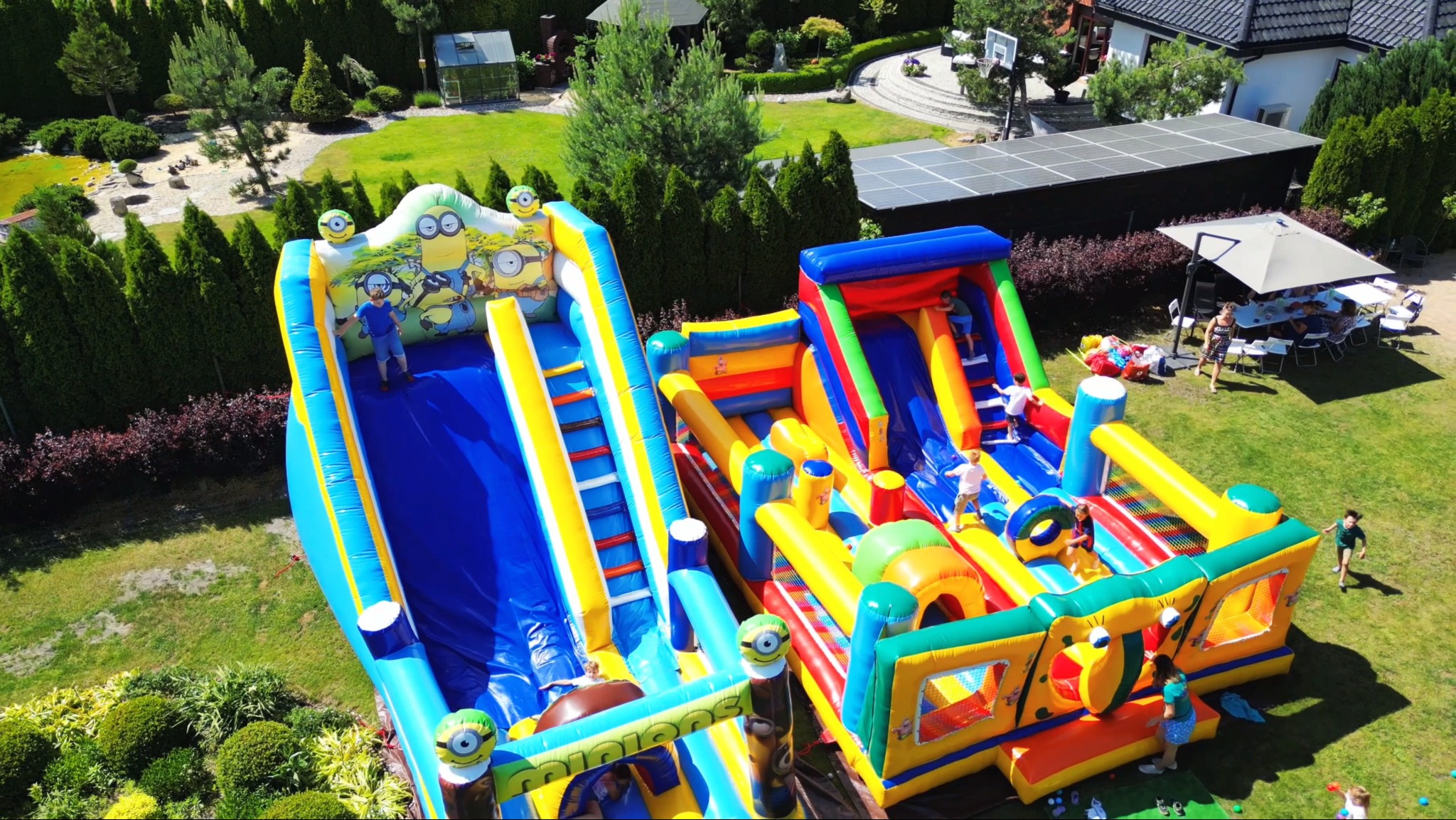 Dwa duże, kolorowe dmuchańce z motywem Minionków na zielonym trawniku, dzieci bawiące się na konstrukcji, w tle dom z panelami słonecznymi i grupa osób siedzących przy stole pod parasolem.