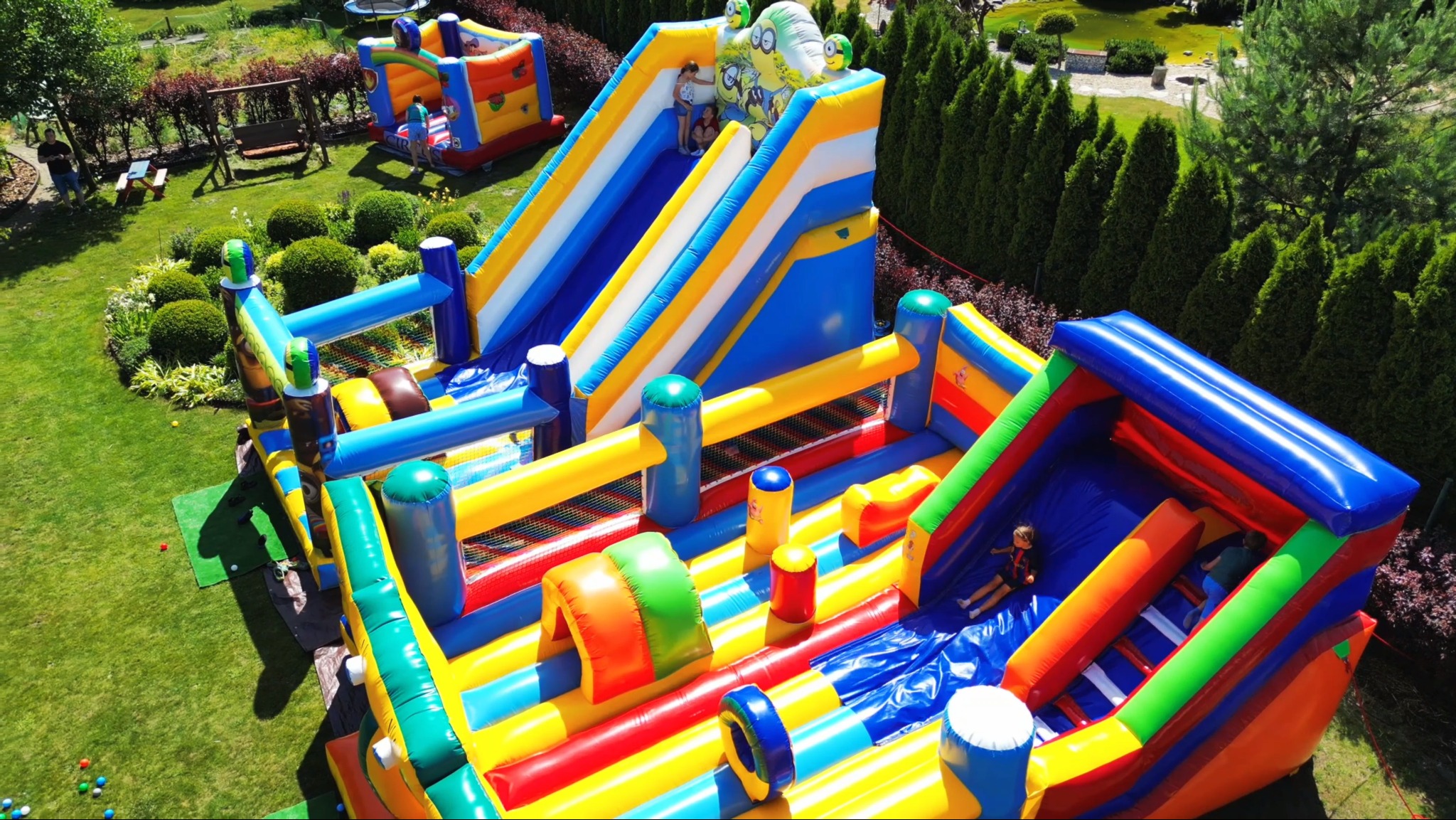 Kolorowy, dmuchany plac zabaw z dwiema zjeżdżalniami i tunelem na zielonym trawniku, z dziećmi bawiącymi się na zjeżdżalniach, otoczony zielenią.
