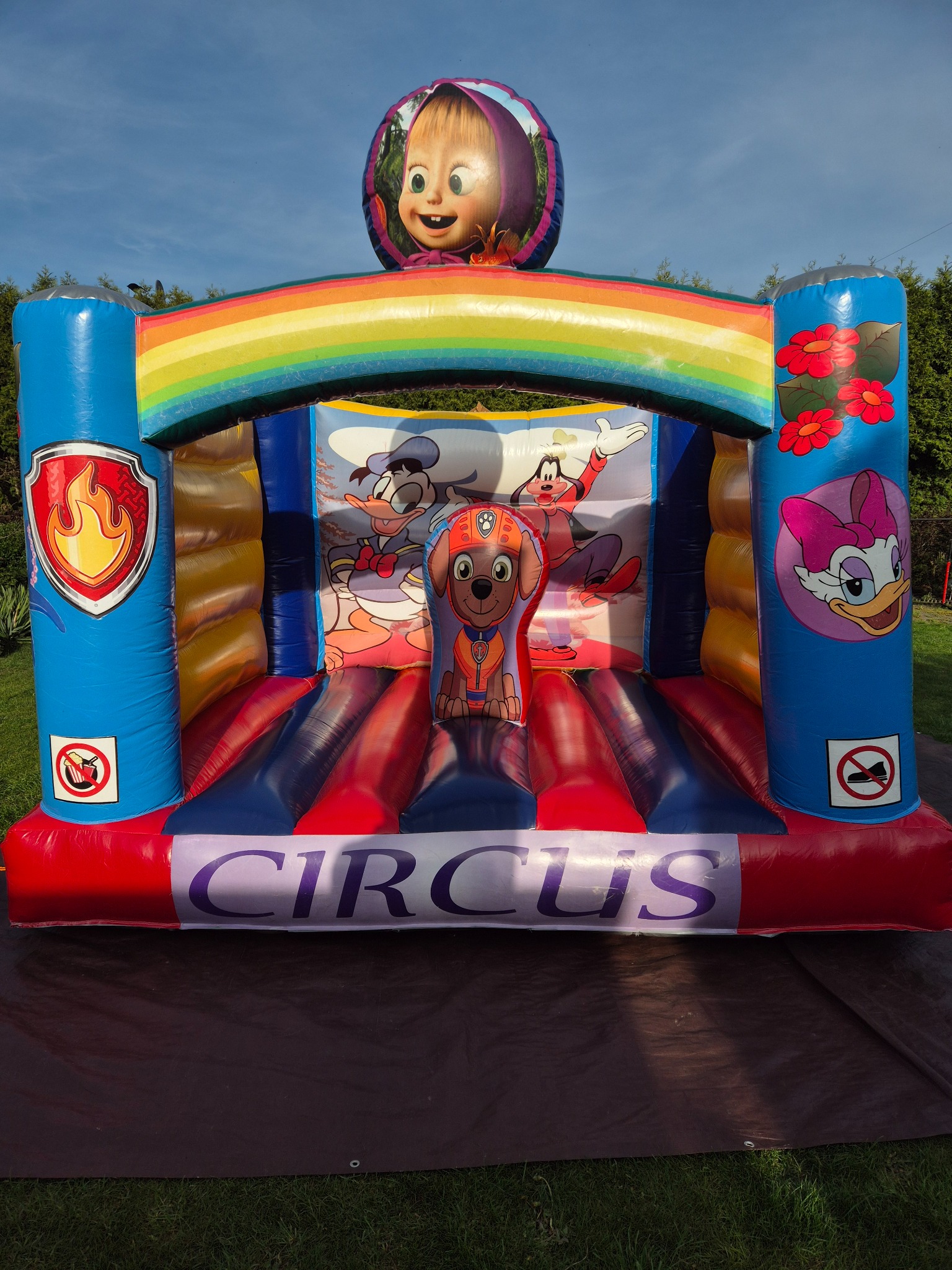 Kolorowy, dmuchany zamek z motywami z bajek, z napisem 'CIRCUS' na dole i wizerunkiem postaci z bajki Masza i Niedźwiedź na górze, ustawiony na trawniku.