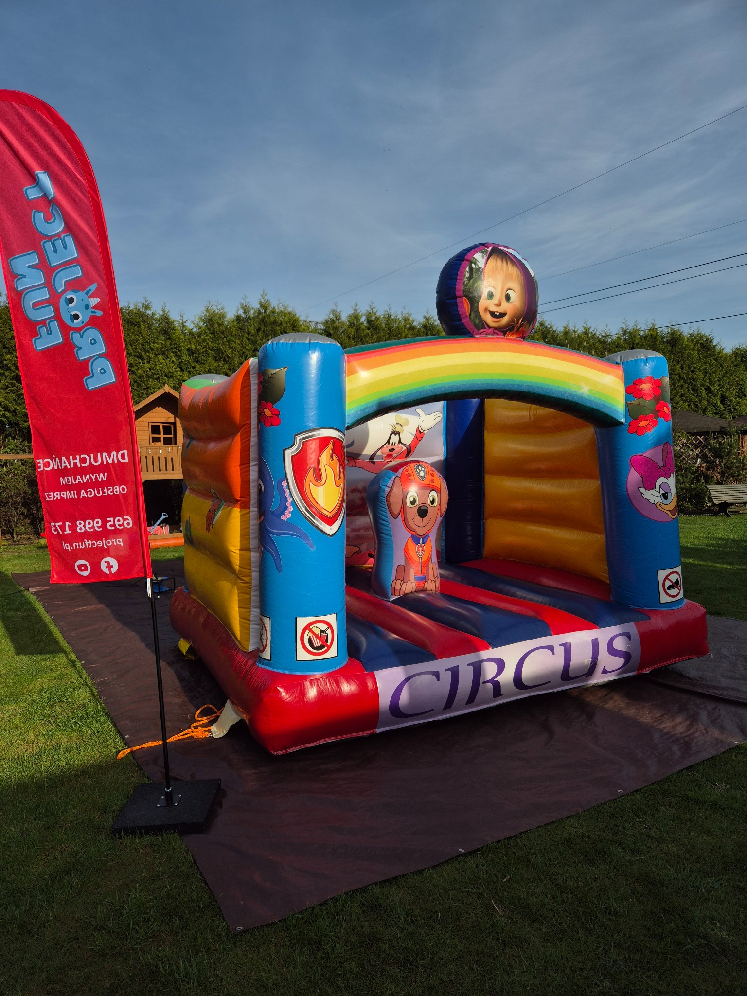 Kolorowy dmuchany zamek z motywami z bajek, ustawiony na trawie obok czerwonego baneru z nazwą firmy, w tle widoczny drewniany domek dla dzieci.