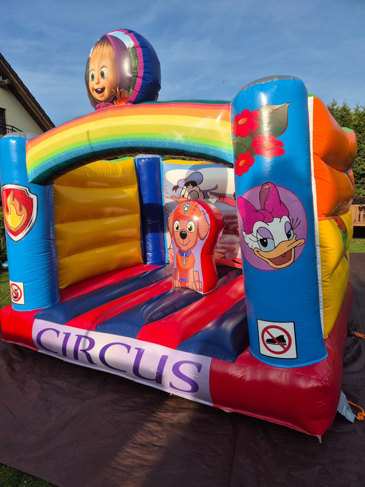 Wielobarwny dmuchany zamek z motywami z bajek, w tym postać Masha na szczycie, Daisy Duck i piesek z Psiego Patrolu, ustawiony na trawniku przed domem.