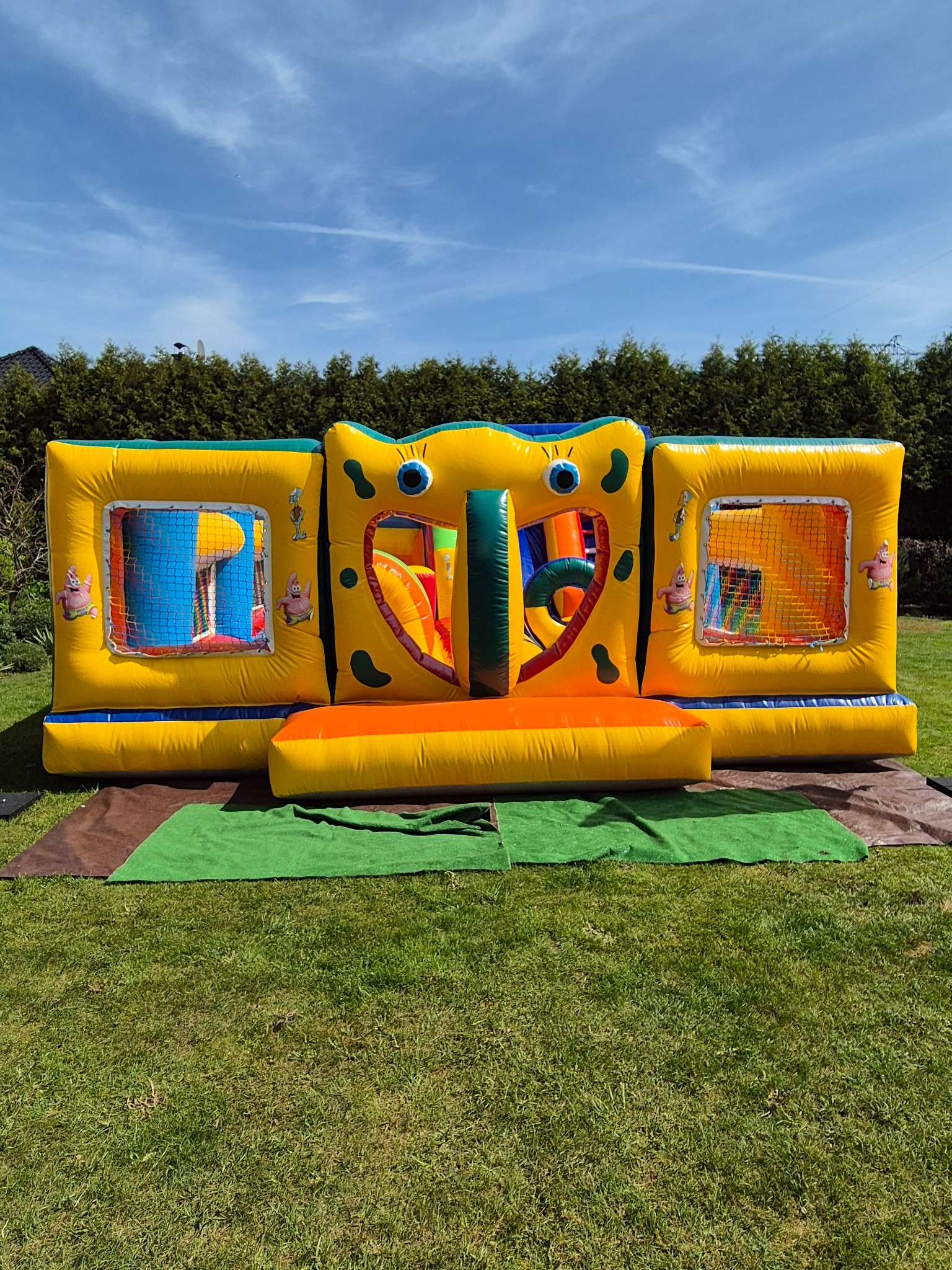 Żółto-pomarańczowy dmuchany zamek z motywami SpongeBoba, ustawiony na trawniku przed żywopłotem, z widocznymi elementami do wspinaczki i zjeżdżania.