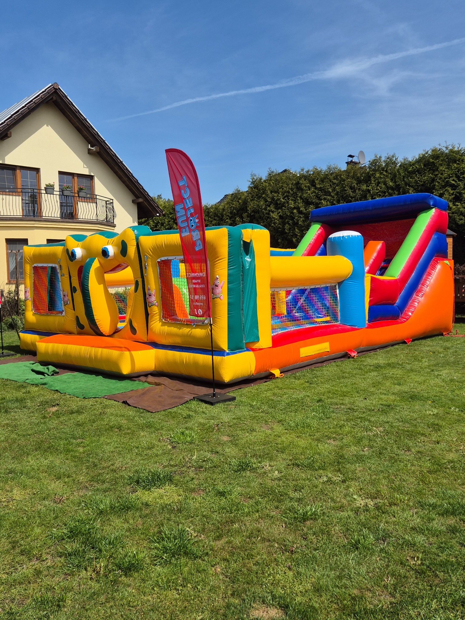 Wielobarwny dmuchany plac zabaw z motywem SpongeBoba na trawniku przed domem z balkonem, widoczny baner reklamowy.