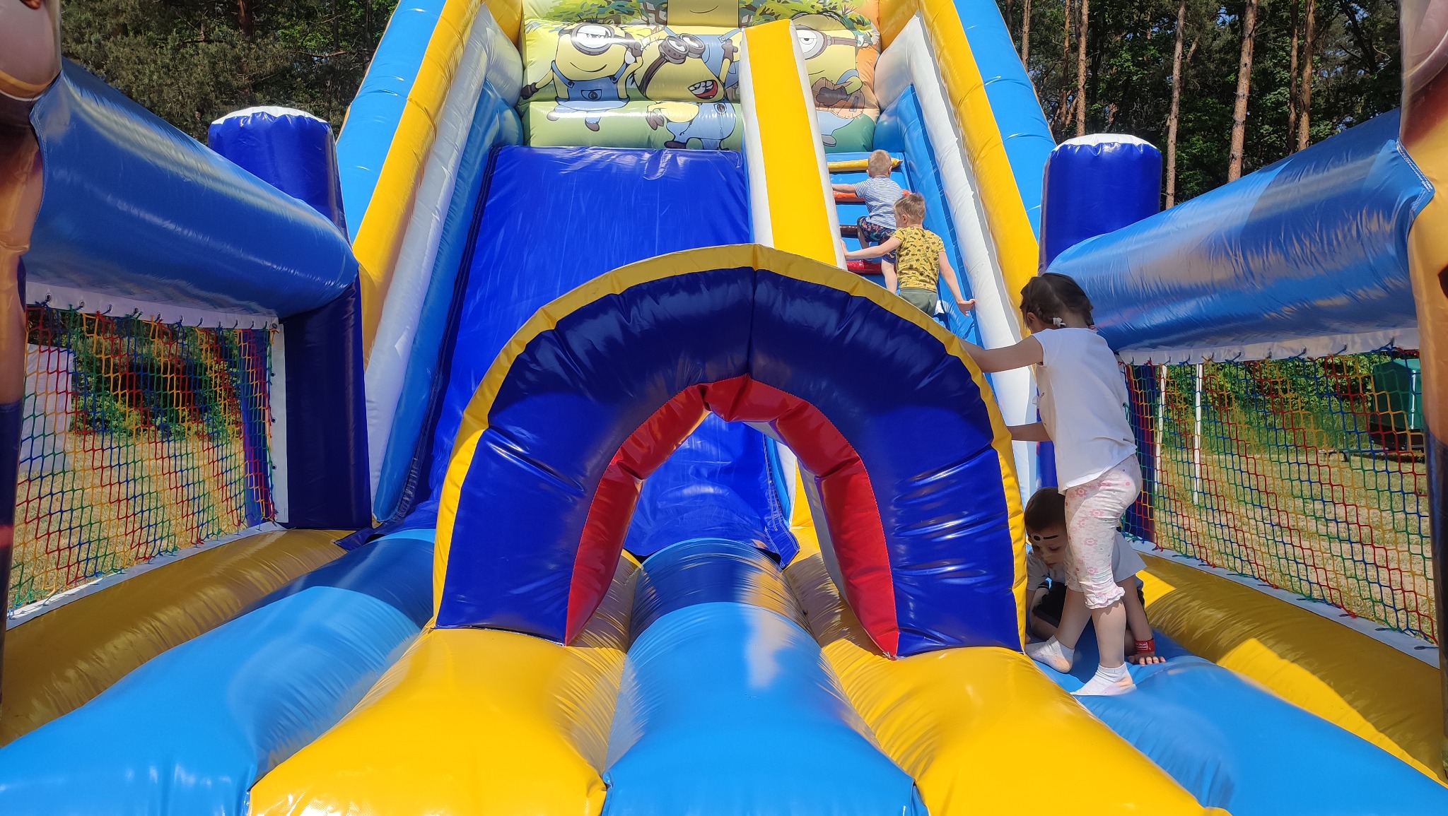 Dzieci wchodzą na dmuchaną zjeżdżalnię z motywem Minionków, widoczna siatka zabezpieczająca i kolorowe elementy konstrukcji.