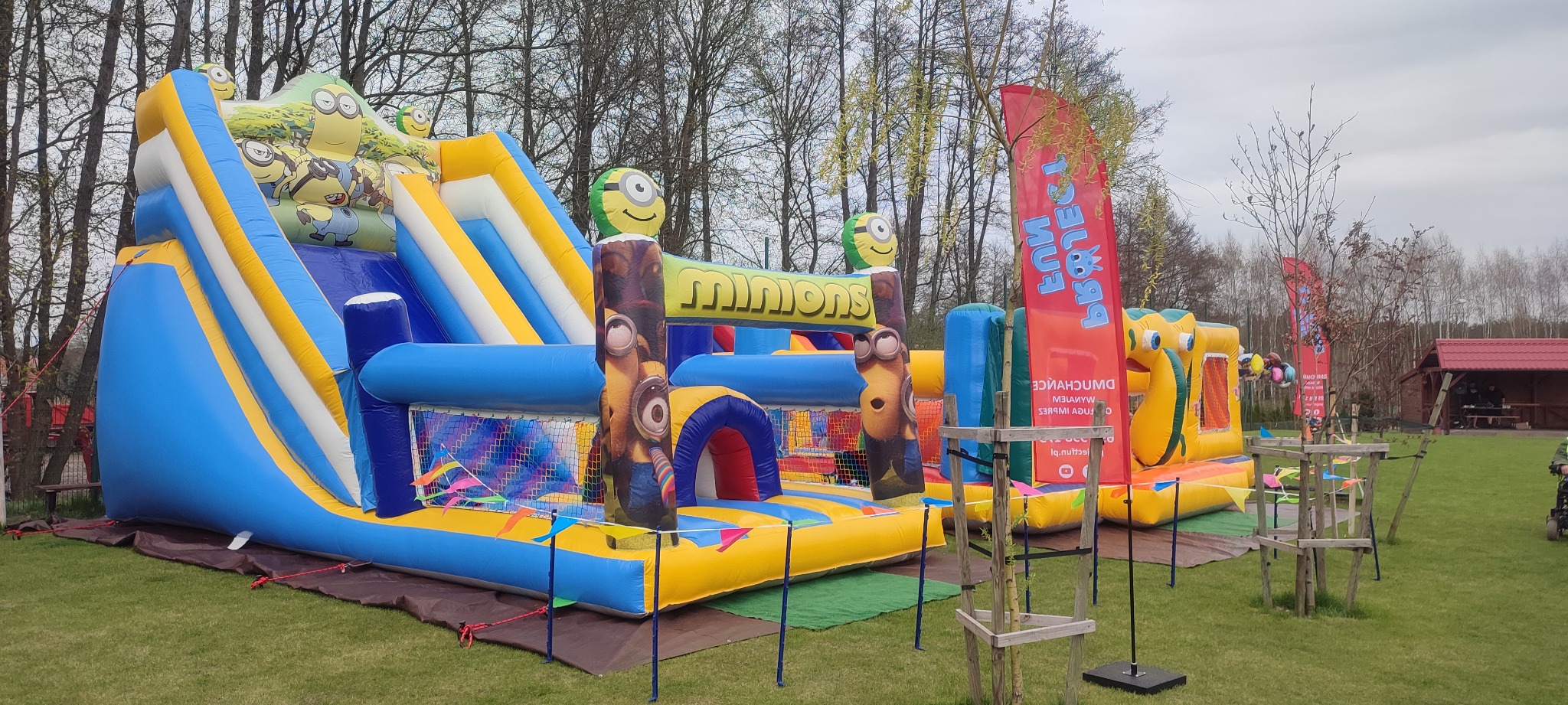 Duży, kolorowy dmuchany plac zabaw z motywem Minionków, zjeżdżalnie i przeszkody, ustawiony na trawiastym terenie zewnętrznym w pobliżu drzew i drewnianej altany.