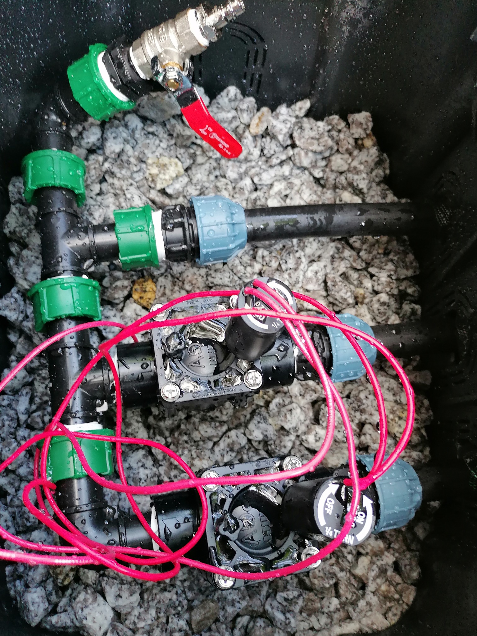 Komponenty systemu nawadniania w czarnej skrzynce wypełnionej drobnymi kamieniami: zawór z czerwoną rączką, czarne rury z zielonymi i niebieskimi złączkami, elektrozawory z czerwonymi kablami.