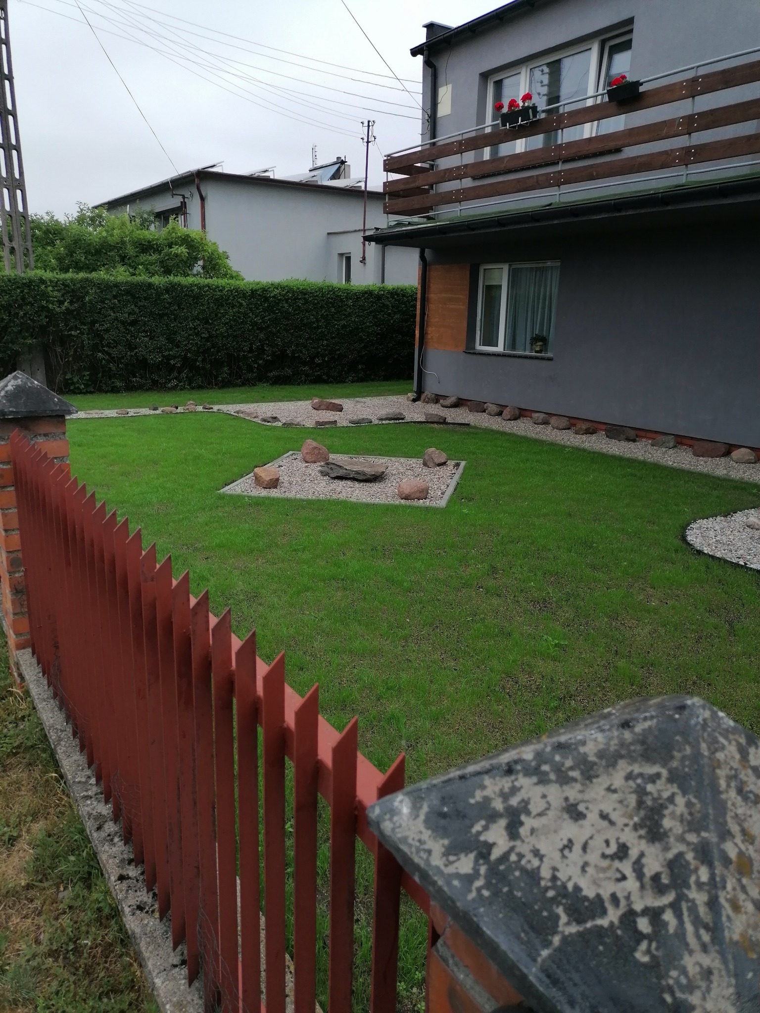 Zadbany trawnik z dekoracyjnymi kamieniami i ścieżką z grysu, widok zza czerwonego, metalowego ogrodzenia.