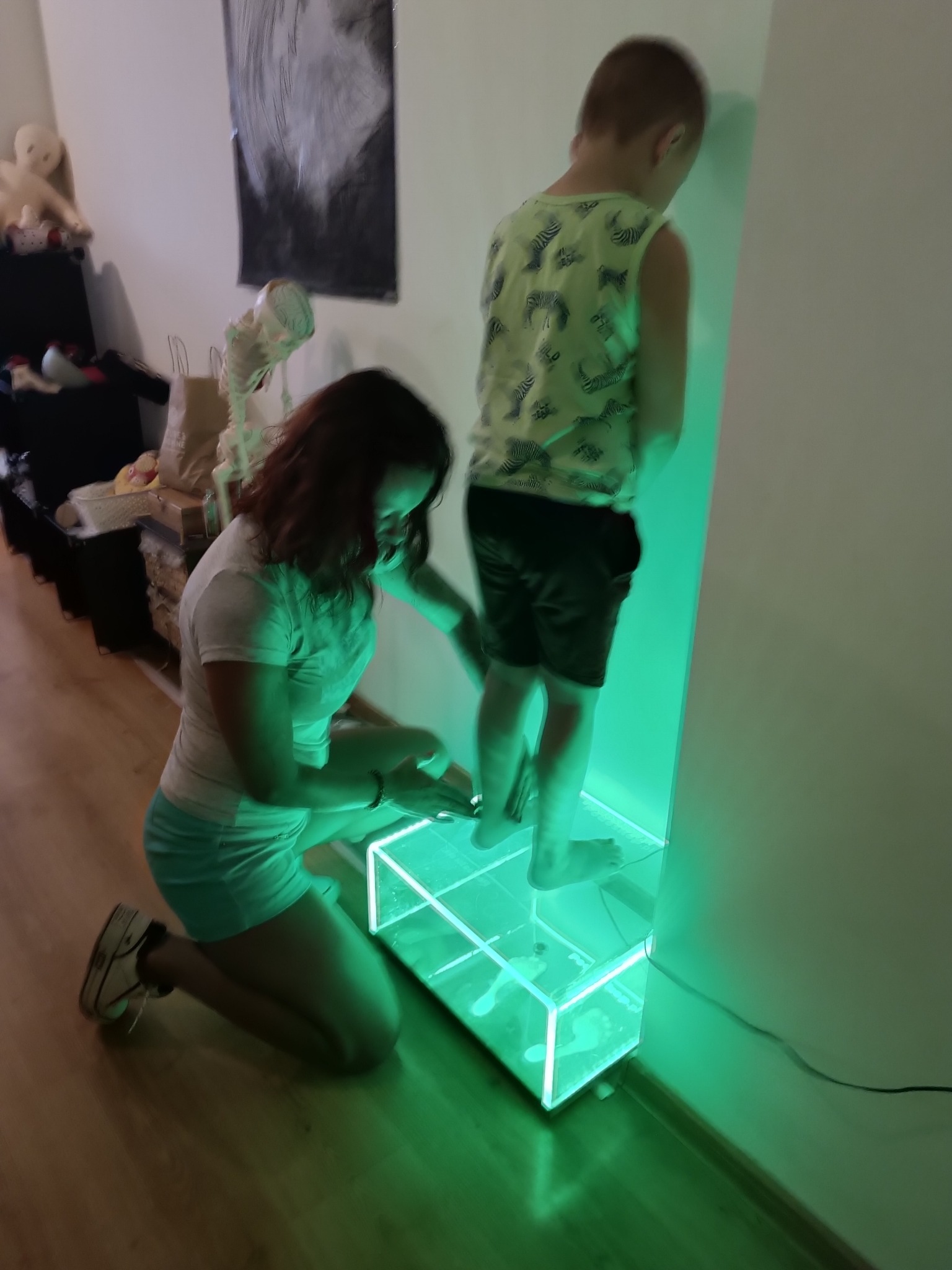 Kobieta na kolanach przy podświetlonym na zielono urządzeniu do badania stóp dziecka, widoczne odciski stóp na szkle.