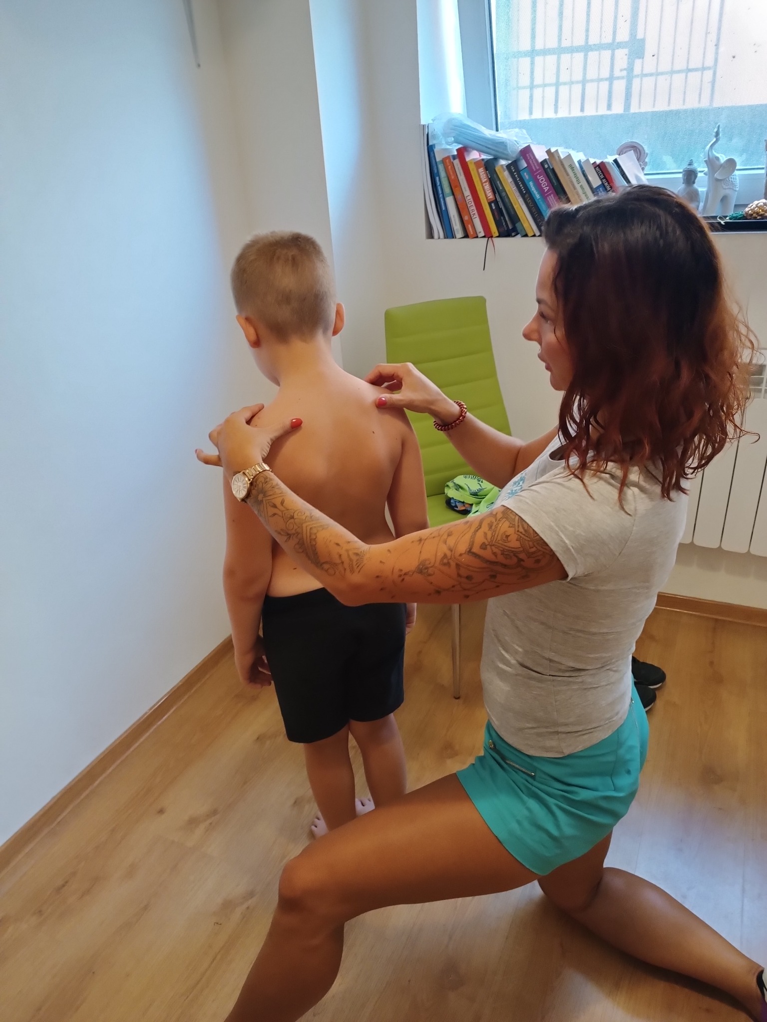 Kobieta z tatuażem na ramieniu, w krótkich turkusowych spodenkach, klęczy i bada plecy dziecka w jasnym pomieszczeniu z drewnianą podłogą, widoczne książki i dekoracje na parapecie okna.