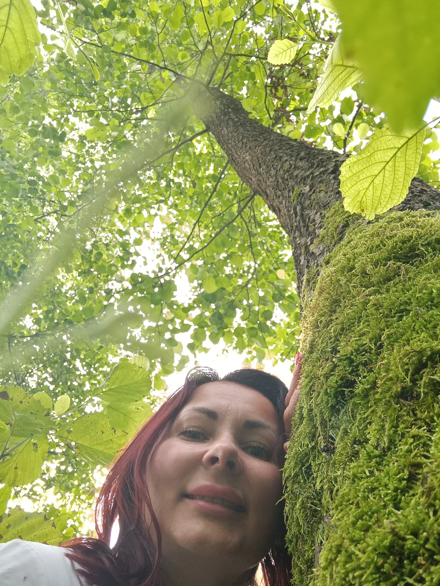 Kobieta opiera głowę o drzewo porośnięte mchem, spoglądając w górę na korony drzew i liście.