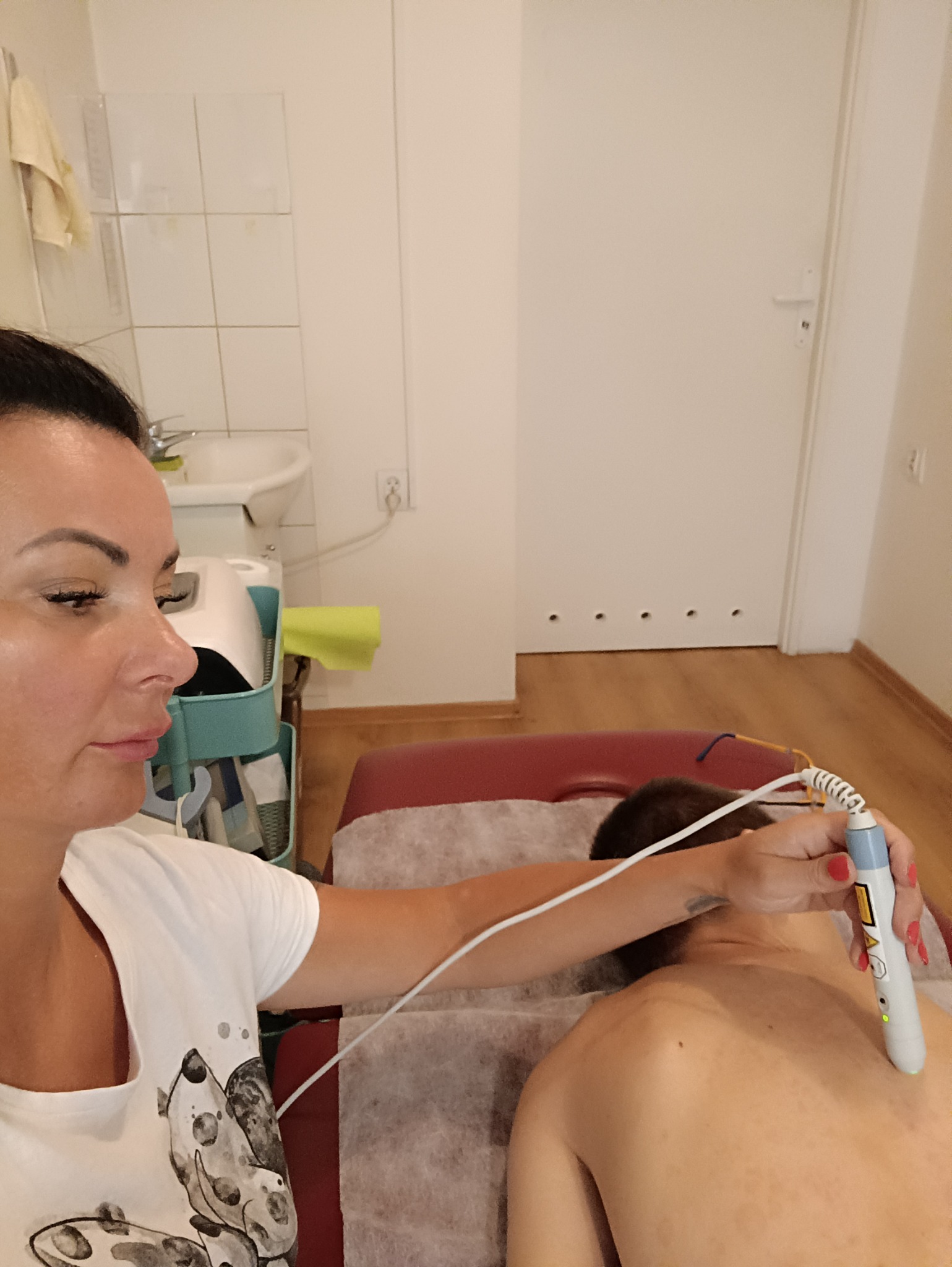 Kobieta w białej koszulce aplikuje urządzenie z przewodem na plecy pacjenta leżącego na czerwonym łóżku w gabinecie.