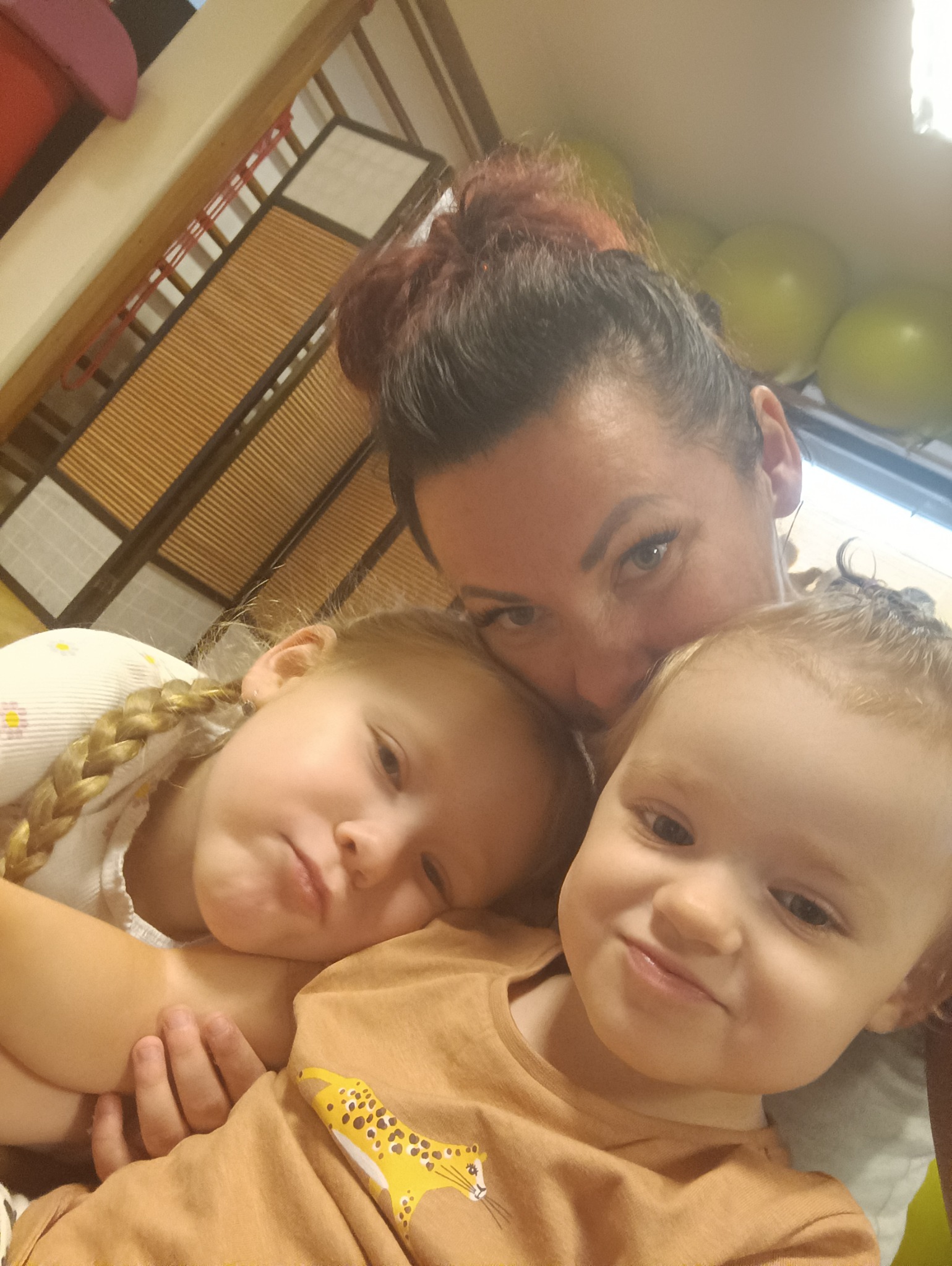 Kobieta z rudymi włosami w koku oraz dwie dziewczynki przytulają się do siebie, widoczne w kadrze od góry, w tle widoczne elementy wystroju wnętrza.