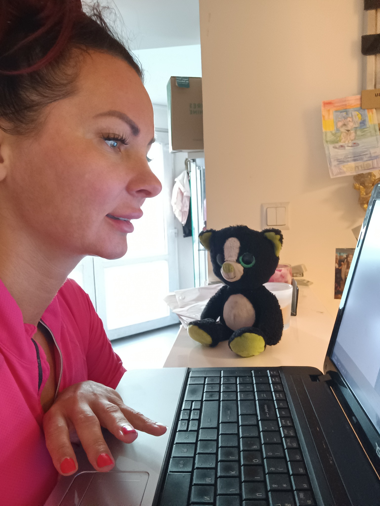 Kobieta w różowej bluzce pisze na laptopie, obok siedzi pluszowy kot z dużymi oczami.