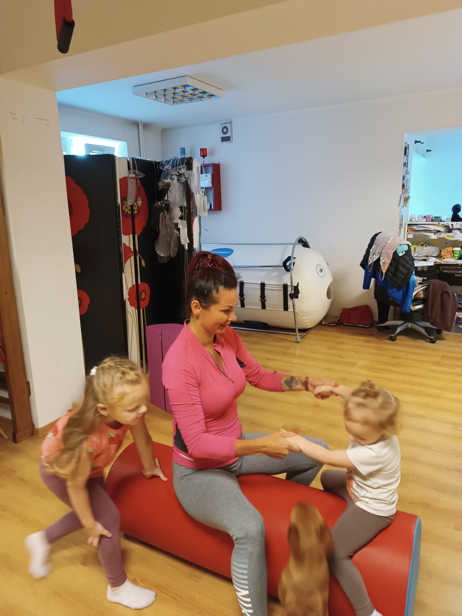 Kobieta w różowej bluzce i szarych legginsach siedzi na czerwonym wałku, trzymając za ręce dwie małe dziewczynki, w tle widoczny sprzęt rehabilitacyjny i biała komora hiperbaryczna.