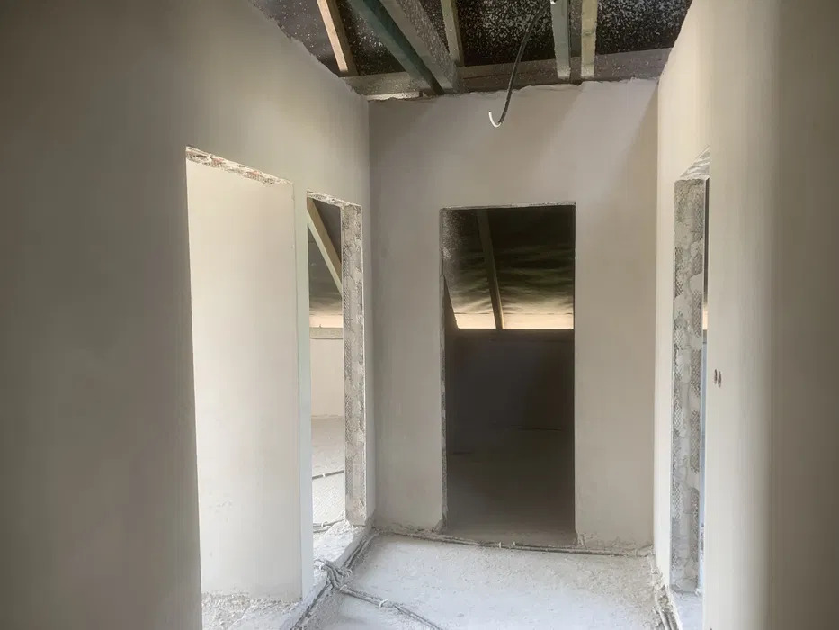 Wnętrze w trakcie prac wykończeniowych, świeżo położone tynki maszynowe na ścianach i suficie poddasza, widoczne instalacje elektryczne przy podłodze oraz surowa konstrukcja dachu.