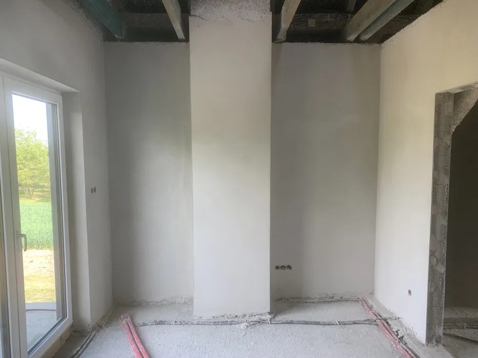 Surowe wnętrze po nałożeniu tynków maszynowych, widoczne instalacje elektryczne w podłodze oraz surowy strop. Świeże tynki na ścianach, widoczne niedoskonałości przy podłodze.