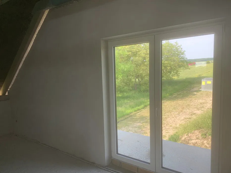Pokój w trakcie wykańczania: surowe tynki na ścianach, widok na zielony krajobraz przez białe, przesuwne okno balkonowe.