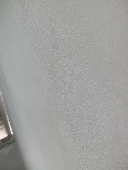 Gładka, jasnoszara ściana z delikatną, równomierną fakturą tynku maszynowego, oświetlona naturalnym światłem z lewej strony.